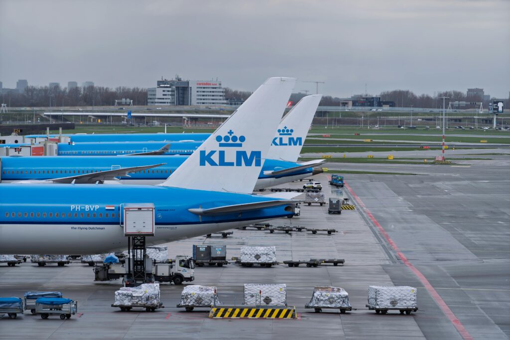 pexels-photo-36581523-36581523 Row of KLM airplanes parked at an airport tarmac with cargo carts, under an overcast sky.